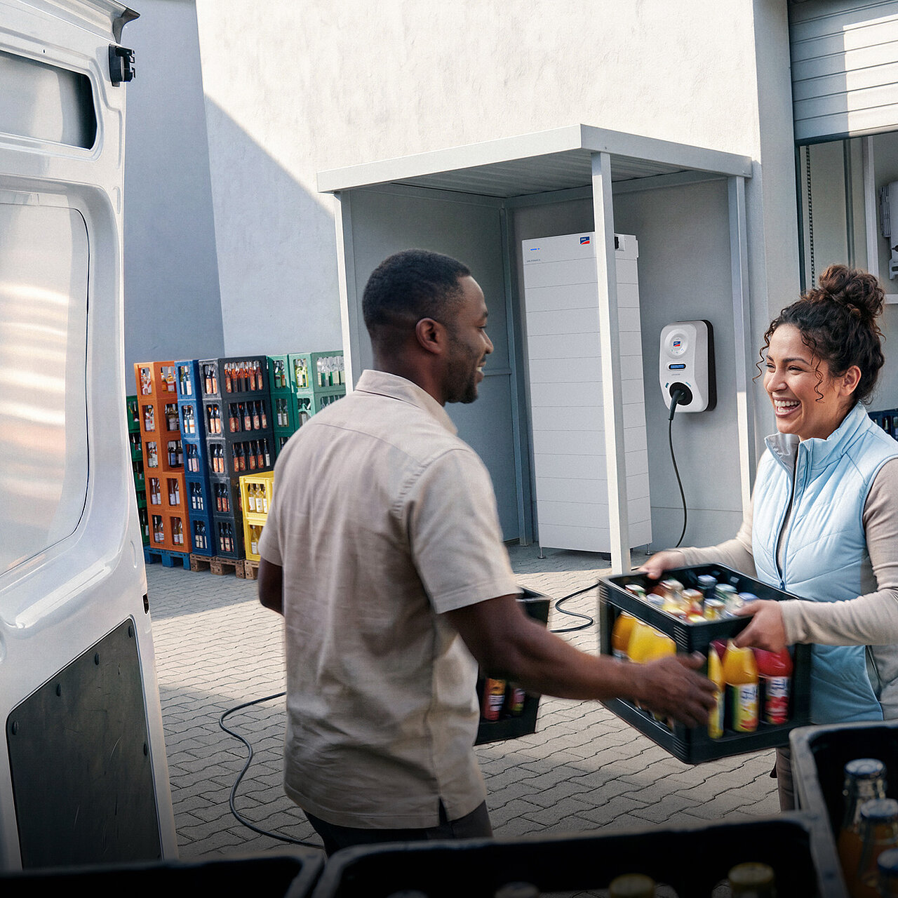 Two people are loading crates of beverages into a delivery van; in the background stands an SMA Storage M, and next to it on the wall hangs an SMA eCharger; a charging cable runs from the SMA eCharger to the delivery van.