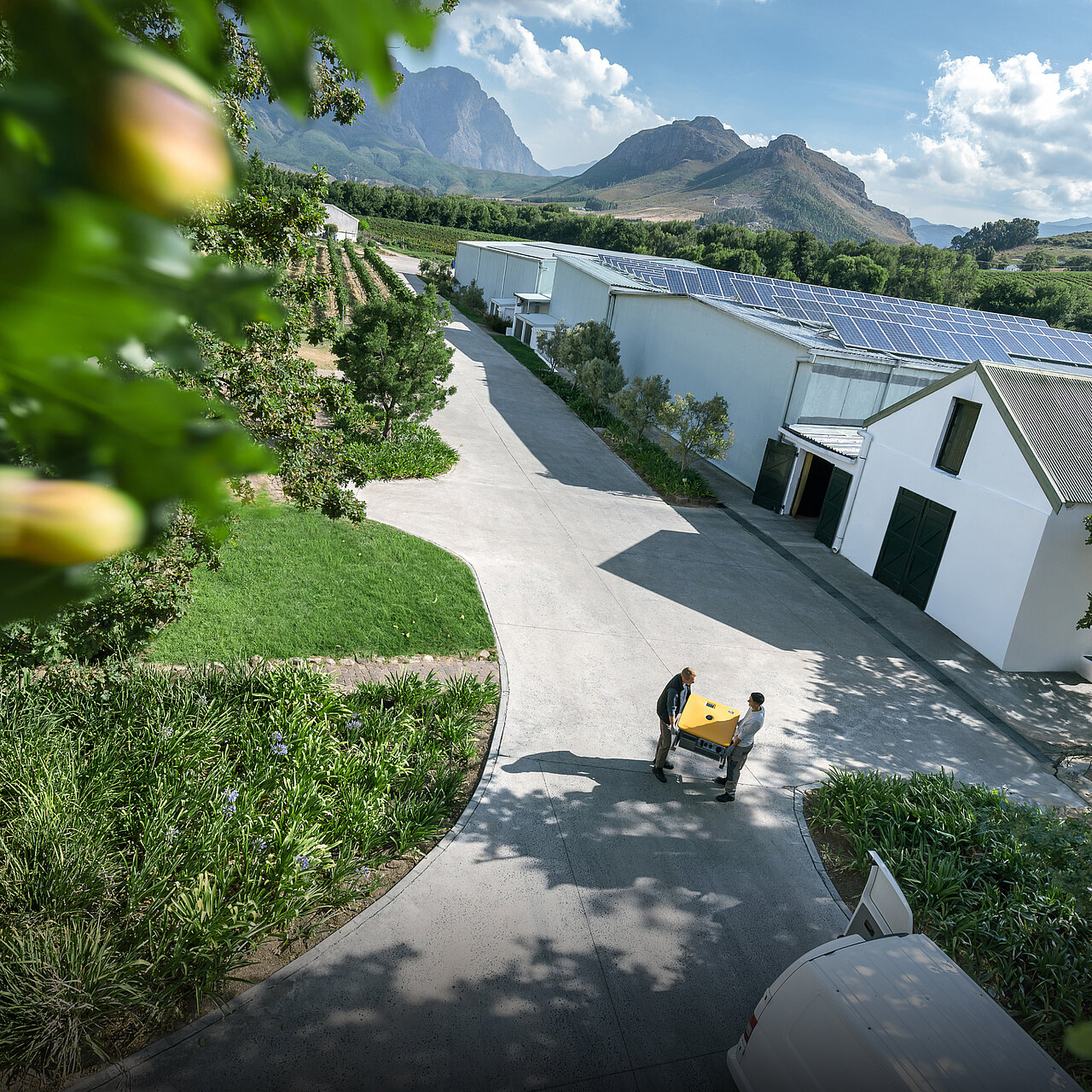 Warehouses with a solar array on the roof; in front of them, two installers are carrying a Sunny Island X.
